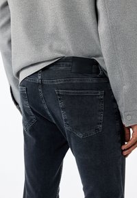 Vaqueros de mezclilla oscura con un diseño ajustado, textura ligeramente desgastada y un parche de cuero negro liso en la parte posterior de la cinturilla.