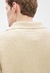 Béžový vlnený sveter s jemnou textúrou, s klopovým golierom a štítkom s logom "Calvin Klein" na zadnej strane.