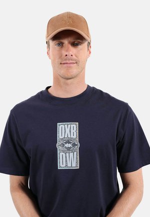 Homme portant une casquette de baseball beige et un t-shirt bleu marine avec un texte graphique blanc central "DXB OW" sur un fond uni.