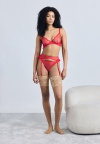 Σετ κόκκινου δαντελένιου εσώρουχου με σουτιέν με ενίσχυση, ζώνη για καλτσοδέτες και διαφανές στρινγκ. Το μοντέλο φοράει μπεζ δαντελένιες κάλτσες με ανθισμένα σχέδια. Απαλό φόντο.