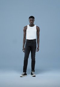 Witte mouwloze tanktop, zwarte skinny jeans en zwarte sneakers. Model staat tegen een lichtblauwe achtergrond. Simpele zilveren ketting zichtbaar.