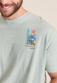 Lichtgroen kortarm shirt van een gestructureerde stof. Heeft een kleurrijk gedrukt ontwerp met palmbomen en een zon, gelabeld "Italiaanse Rivièra."