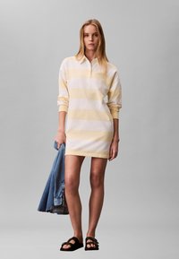 Femme portant une robe à manches longues rayée jaune et blanche, tenant une veste en jean bleue et des sandales noires, sur un fond gris uni.