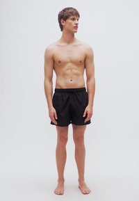 Μαύρα μαγιό σορτς με ελαστική μέση και κορδόνι, φορεμένα από μοντέλο που στέκεται ξυπόλυτο μπροστά σε λευκό φόντο.