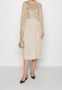 Gouden pailletten top met lange mouwen, gecombineerd met een geplooide beige midi-jurk. Het model houdt een kleine crèmekleurige handtas vast en draagt zwarte puntige hakken.