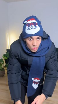 Chapeau en tricot bleu foncé avec motif de Père Noël et lunettes de soleil rouges, assorti à une écharpe. Le modèle porte une doudoune noire.