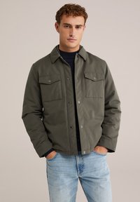 Giovane uomo con capelli ricci che indossa una giacca verde oliva con bottoni sopra una maglietta nera e jeans azzurri, mani nelle tasche, sfondo neutro.