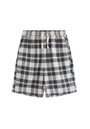 Teenagedreng står barfodet iført en mørkeblå t-shirt over en lyseblå skjorte og ternede shorts mod en ensfarvet hvid baggrund.