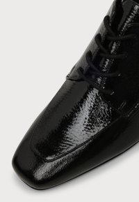 Chaussure habillée en cuir verni noir brillant avec embout carré et lacets noirs, présentant une surface texturée et réfléchissante.