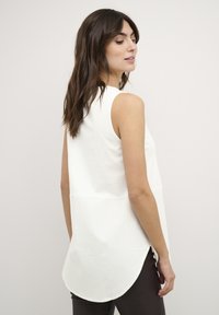Top bianco senza maniche con scollatura rotonda, caratterizzato da uno spacco posteriore per una buona circolazione dell'aria e una texture liscia. Abbinato a leggings scuri con motivo.