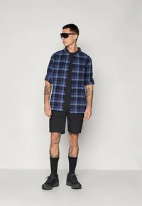 Sjekket blå og svart skjorte, svart t-skjorte, svarte shorts, svarte sokker, svarte joggesko og solbriller. Rene linjer og avslappet passform.