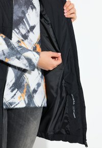 Svart jacka med dragkedja, som visar ett slätt, glänsande inre och en etikett. Bärs över en mönstrad, mjuk tröja med orange detaljer.