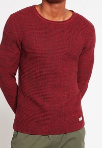 Punainen teksturoitu pitkähihainen villapaita, jossa on pyöreä pääntie ja ribbikuvio. Sisältää pienen logotagin alahelassa.