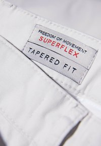 White tapered fit label with red text stating "SUPERFLEX” and "FREEDOM OF MOVEMENT." Neat stitching along edges, smooth fabric texture.