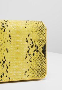Gul pung med sort og mørkebrun slangeskinmønster, tekstureret overflade og afrundede hjørner mod en ensartet lys baggrund.