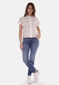 Blusa bianca a maniche corte con stampa floreale rosa, chiusura con bottoni, abbinata a jeans blu e sneakers bianche. Texture liscia, design casual.
