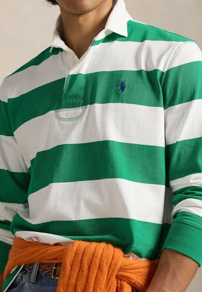 Zöld-fehér csíkos galléros rugby póló, a mellkasán egy kis kék logóval. Narancssárga kötött pulóver van a derékra kötve.
