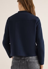 Femme aux longs cheveux bruns portant un haut à manches longues bleu marine et un jean noir délavé, vue de dos sur un fond uni.