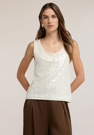 Joven con cabello largo y castaño lleva una blusa blanca sin mangas con lentejuelas y pantalones anchos marrones sobre un fondo liso.