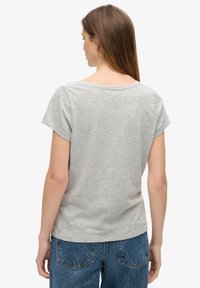 Kvinde med langt brunt hår set bagfra iført en lysegrå kortærmet top og blå denimjeans mod en hvid baggrund.