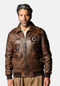 Veste en cuir brun avec fermeture éclair à l'avant, poignets côtelés, deux poches avant et plusieurs patchs brodés. Texture lisse et design classique.