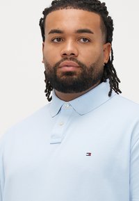 Lyseblå poloshirt lavet af tekstureret stof med en tre-knaps lukning og en lille logo nær brystet.