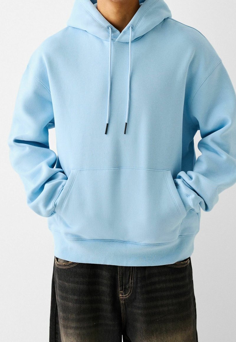 Personne portant un sweat à capuche bleu clair avec poche avant et cordons, associé à un jean noir délavé.