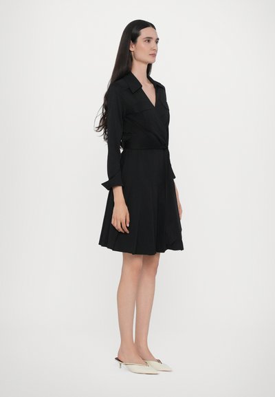 Čierne šaty s dlhými rukávmi a výstrihom do V, s priliehavým pásom a plisovanou sukňou. Modelka má na sebe krémové open-toe mule topánky.