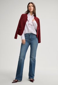 Rode textuurt blazer over een witte blouse met een strik, gecombineerd met lichtblauwe flared jeans en bordeauxrode puntige schoenen. Neutrale achtergrond.