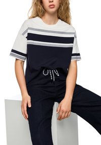 Navy and white striped oversized T-shirt with short sleeves, featuring a drawstring waist and soft fabric. Worn with dark blue jeans.