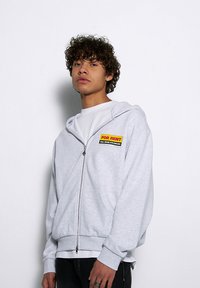 Szara bluza z kapturem na zamek, wykonana z polaru, z wyszywaną łatą z przodu z żółtym i czarnym napisem. Kieszenie i ściągacze na rękawach dopełniają projekt.