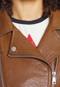 Veste de moto en cuir marron avec des fermetures éclair argentées, des boutons-pression et une finition texturée. Portée par-dessus une chemise blanche avec un motif graphique rouge.