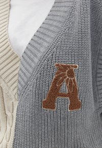 Cardigan lavorato a maglia grigio e beige con una toppa testurizzata a forma di "A" marrone sul lato grigio, caratterizzato da distinti motivi a costine e una texture morbida.