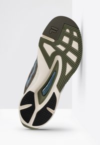 Suola di sneaker composta da una miscela di materiali in gomma verde scuro, beige e nera, con motivi testurizzati e un elemento di ammortizzazione in gel visibile.