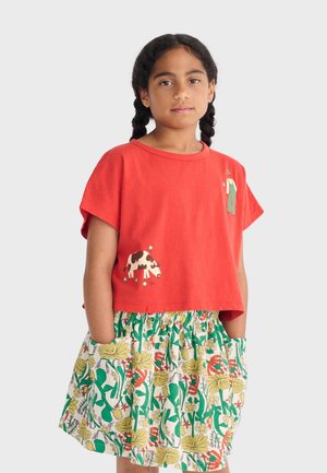 Fille aux cheveux tressés portant un t-shirt rouge court avec des motifs de vache et de personne, ainsi qu'une jupe à motif floral avec des poches, debout devant un fond blanc.