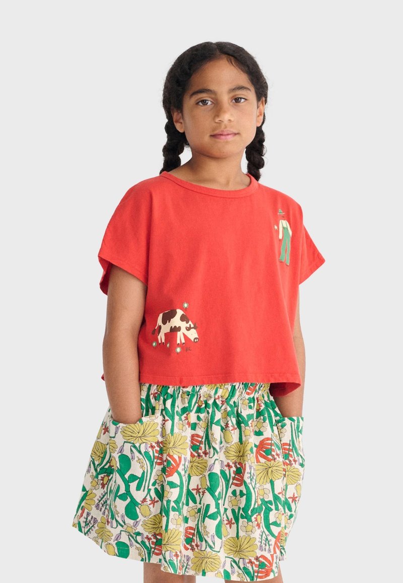 Mädchen mit geflochtenem Haar trägt ein rotes, kurzes T-Shirt mit Kuh- und Personenmotiven sowie einen geblümten Rock mit Taschen und steht vor weißem Hintergrund.