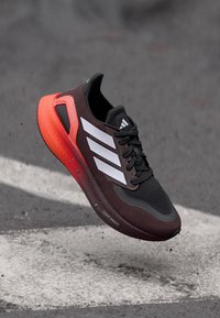 Zwarte en kastanjebruine hardloopschoen met witte strepen, een gestructureerde bovenkant en een feloranje zool. Heeft "Light Boost" branding aan de zijkant.