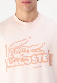 Hellrosa Sweatshirt mit einem großen, verblassten korallenfarbenen Krokodil-Logo und dem Text "LACOSTE". Weiches Baumwollmaterial mit klassischem Rundhalsausschnitt.