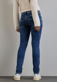 Kapeat denim-farkut tummansinisessä pesussa, jossa on sileä pinta. Ominaisuuksina perinteiset takataskut ja kuluneet reunat nilkoissa. Valaistuna valkoisilla lenkkareilla.