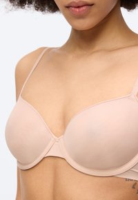 Γυναίκα που φοράει σουτιέν σε γυμνή απόχρωση, με ελαφριά επένδυση και λεπτές τιράντες, εμφανιζόμενη από τους ώμους έως το μέσο του κορμού σε απλό φόντο.