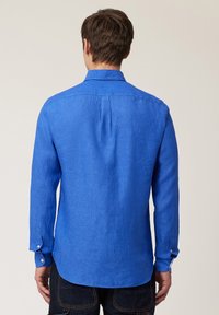 Camisa de manga larga para hombre en lino azul vibrante, con pliegue en la espalda, dobladillo redondeado y puños abotonados. Textura suave y ajuste relajado.