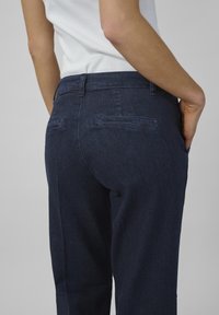 Een persoon in een aansluitend wit topje en donkerblauwe broek staat met de rug naar de camera, handen in de zakken, en toont het ontwerp van het kledingstuk.