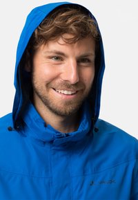 Blaue Kapuzenjacke mit strukturiertem Finish, ausgestattet mit einem Reißverschluss an der Front, verstellbaren Kordelzügen und Branding auf der linken Seite.