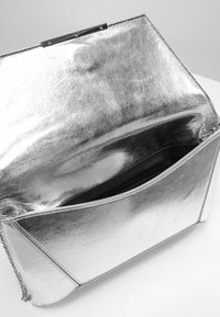 Silverfärgad metallisk clutchväska med kedjehandtag, visad öppen för att avslöja ett svart blixtlåsfack inuti.