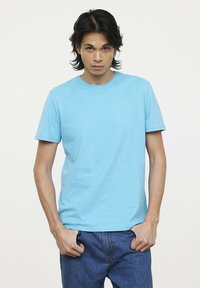 Himmelblaues Baumwoll-T-Shirt mit kurzen Ärmeln und Rundhalsausschnitt. Mit einem kleinen, geprägten Logo auf der linken Brust. Getragen mit blauen Jeans.