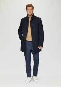 Marineblauwe wollen jas met hoge kraag, knoopsluiting en ritszak, gedragen over een beige trui en marineblauwe broek, gecombineerd met witte sneakers.