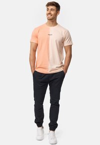Camiseta de manga corta en degradado de melocotón y crema, con un corte ajustado y un pequeño logo en el pecho, combinada con pantalones negros ajustados y zapatillas blancas.