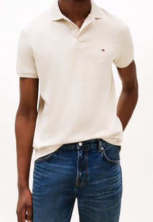 Mand iført en cremefarvet polo med kortærmer med lille logo og blå denimjeans med hænder afslappet langs siden.