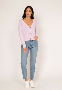 Cardigan lilas côtelé avec cinq boutons marron, décolleté en V, porté sur un jean bleu clair et des baskets blanches. Design simple et décontracté.