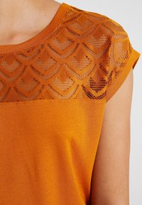 Ärmelloses oranges Oberteil mit transparenter dekorativer Spitze im Schulter- und Brustbereich, die geometrische Muster auf der oberen Brust und den Schultern zeigt.